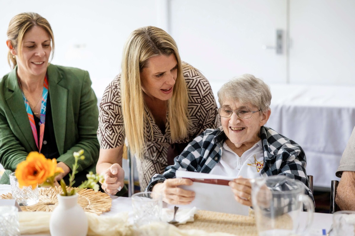 An elderly woman happily reading a document about Support at Home levels, accompanied by a supportive staff member leaning in to help her read and another woman seated nearby.