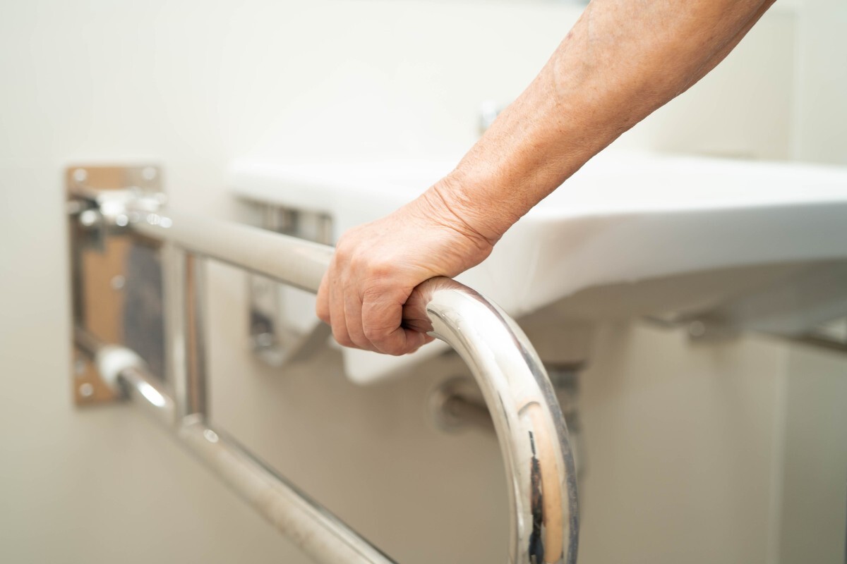 Older person holding a stainless-steel grab rail beside a bathtub.